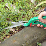 Person using green garden shears to trim plants near a concrete edge.