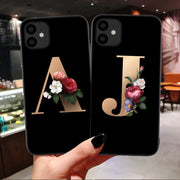 Two black phone cases with floral initials held in a hand against a blurred indoor background.