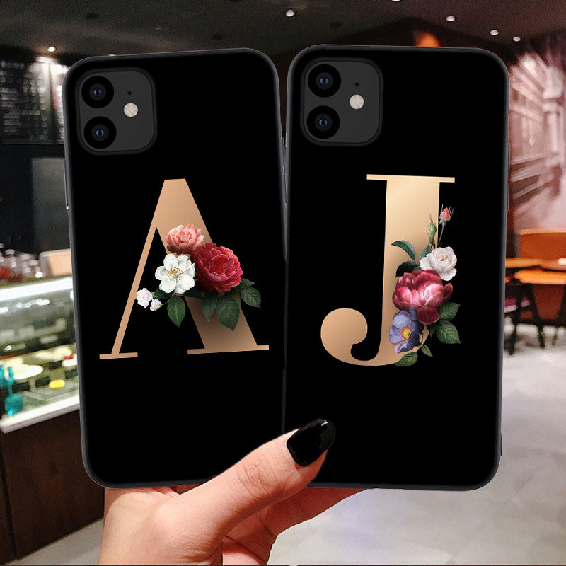 Two black phone cases with floral initials held in a hand against a blurred indoor background.