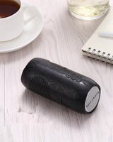 Black portable speaker on a light wooden surface with a cup of tea and notebook in the background.