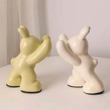 Two ceramic figures, one yellow and one white, on a light surface with a neutral background.