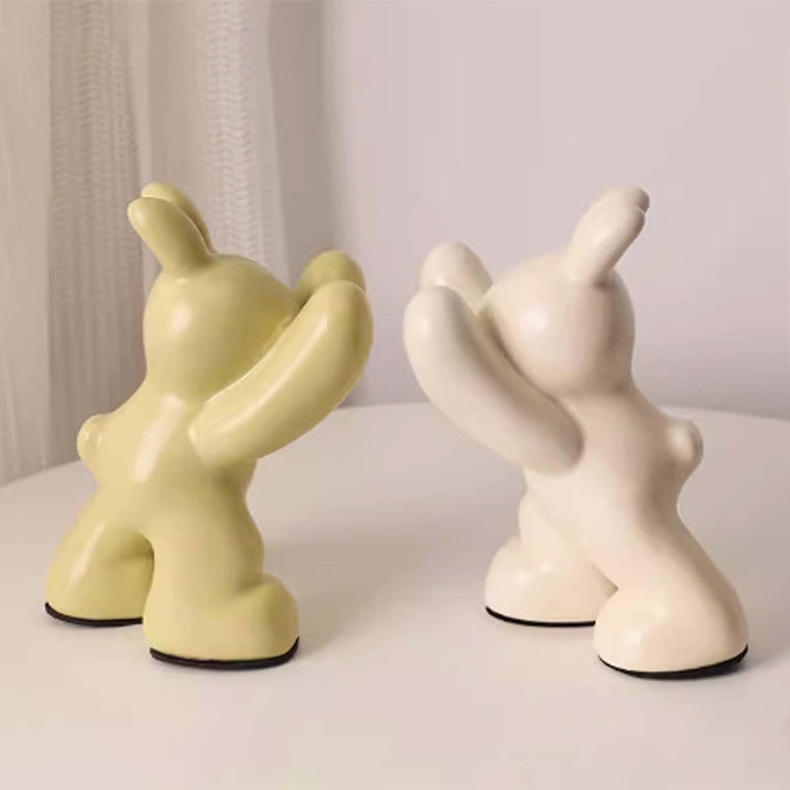Two ceramic figures, one yellow and one white, on a light surface with a neutral background.