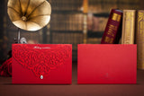 Two red envelopes with intricate designs on a wooden surface with books and a vintage lamp in the background.