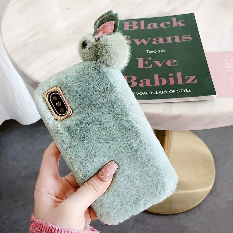 Green fluffy phone case with a bunny ear pom-pom held by a hand, with a book in the background.