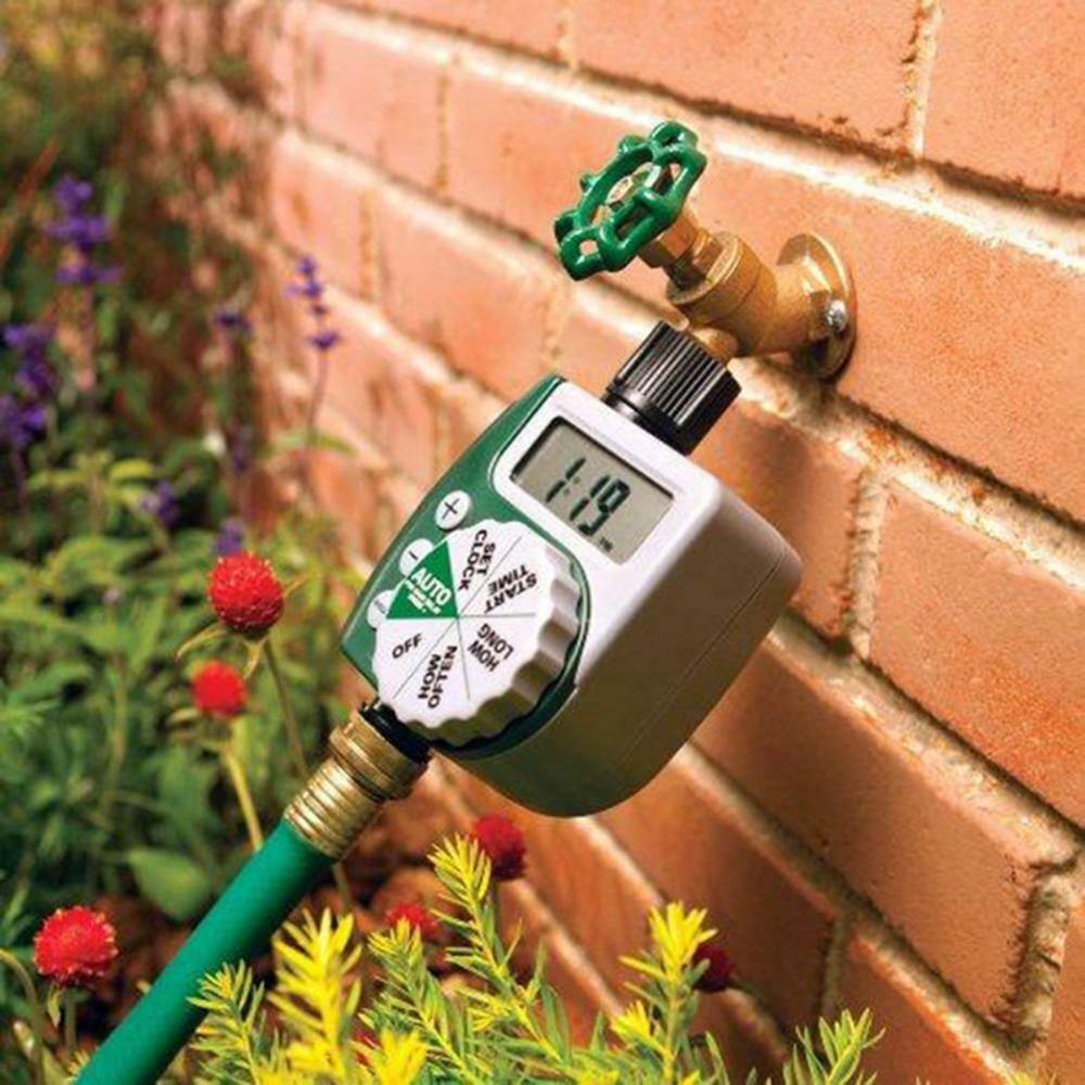 Digital water timer attached to a garden hose with a brick wall and plants in the background