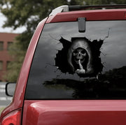 Car window with a skull decal showing a finger to the camera, outdoors.