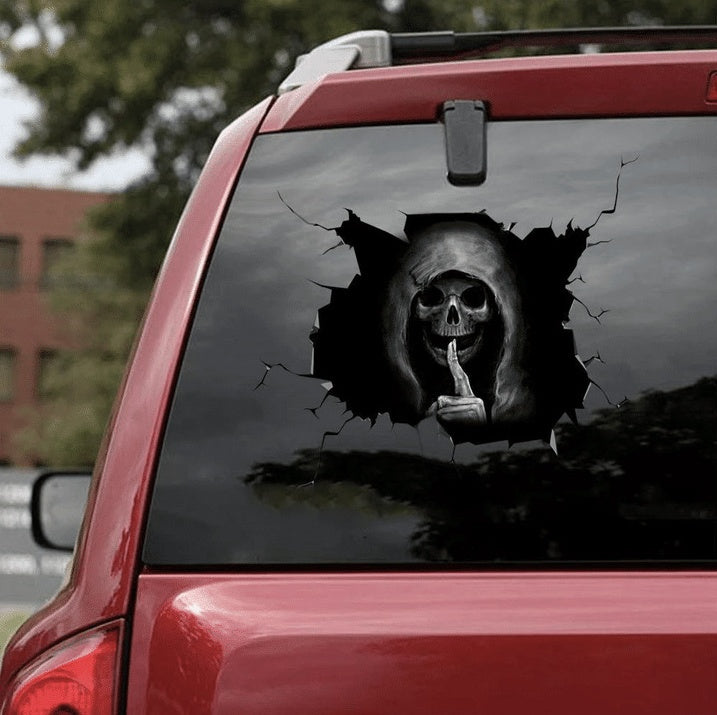 Car window with a skull decal showing a finger to the camera, outdoors.