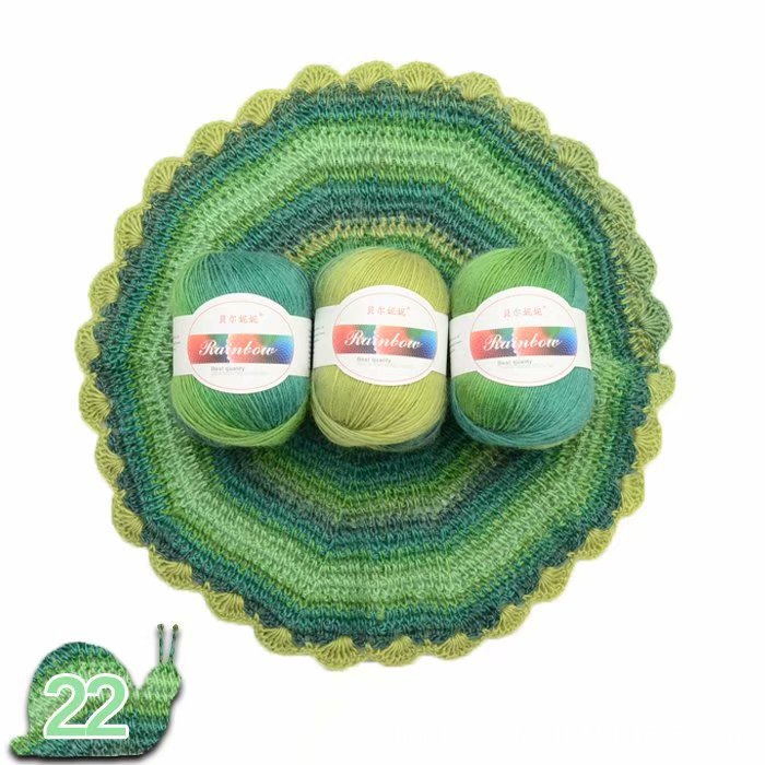 Three balls of green yarn with labels on a crocheted green and blue dish, with a small green crochet hook.