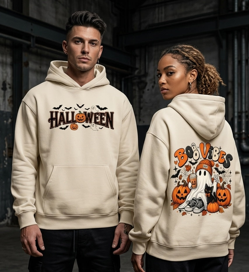 A man and a woman modeling cream-colored hoodies with Halloween graphics. The man wears a front view of the hoodie featuring 'HALLOWEEN' text with pumpkins and bats.