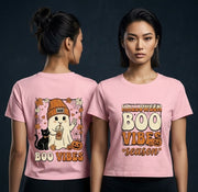 Two women modeling a light pink graphic T-shirt against a dark studio background. The shirt shows a Halloween-themed print: one view has a cartoon ghost, black cat, jack-o'-lantern, and the words 'BOO VIBES'; the front view has 'HALLOWEEN BOO VIBES season' in large cream and orange lettering.