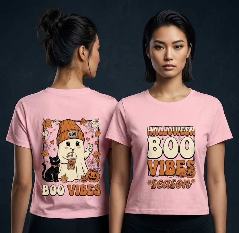 Two women modeling a light pink graphic T-shirt against a dark studio background. The shirt shows a Halloween-themed print: one view has a cartoon ghost, black cat, jack-o'-lantern, and the words 'BOO VIBES'; the front view has 'HALLOWEEN BOO VIBES season' in large cream and orange lettering.
