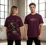 A man and a woman modeling matching maroon short-sleeve t-shirts in a bright, sunlit brick warehouse. The woman shows the back of the shirt, which features a large graphic of a skateboard with skulls and the words 'Skate Board' in white and red, surrounded by small repeated 'Skate Board' logos.