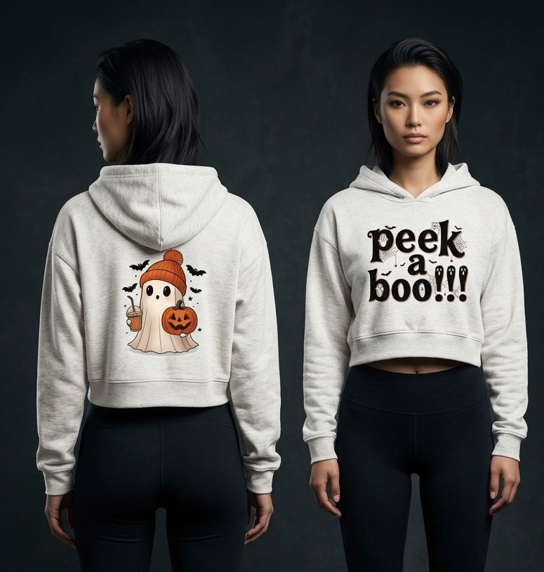 Two models shown against a dark studio background, wearing a cropped white hoodie from the front and back. The front view shows the text 'peek a boo!!!' in black, and the back view shows a cartoon ghost wearing an orange beanie and holding a jack-o'-lantern.